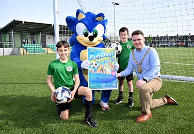 Lord Mayor with two boys in football gear and character Sonic the Hedgehog at Marrowbone Millennium Park football pitch. They hold a poster and footballs promoting a Fun Day event at the park.