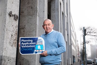 Councillor Arder Carson standing beside an office building holding a sign promoting council's Planning Application Validation Checklist consultation which is now open